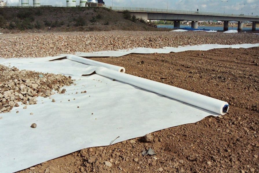 Ausgerollte weiße Geotextilfolie auf einem bräunlichen Schotteruntergrund, mit einer Brücke im Hintergrund
